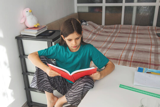 Girl reading red book at home, sitting on desk beside bed, focused on pages, representing childhood education, independent learning, literacy, concentration, homework, knowledge development