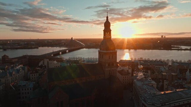 Sun flare behind Riga Dome cathedral tower from drone, Latvia. Aerial side move past the clock tower of Riga, Latvia, as sun flare glows behind it above old town rooftops, river and bridges at sunset.