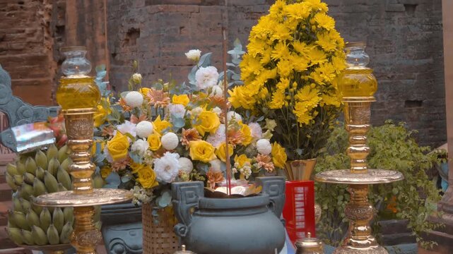 A devotional altar decorated with vibrant a large bunch of bright yellow chrysanthemums or marigolds and white peons and glowing glass oil lamps includes tall, golden-toned glass candle or oil lamps o