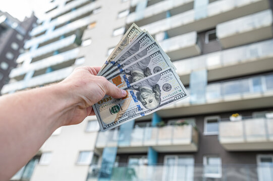 Male Hand Holding Stack of 100 US Dollar Banknotes Against Modern Apartment Buildings Exterior
