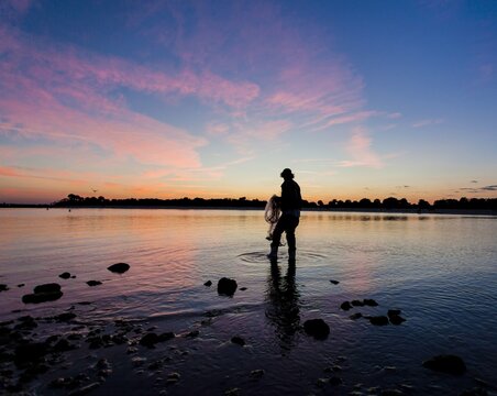 Silhouette of a fisherman holding a cast net standing in the water during a vibrant pink and blue sunset, traditional fishing coastal lifestyle scenic landscape
