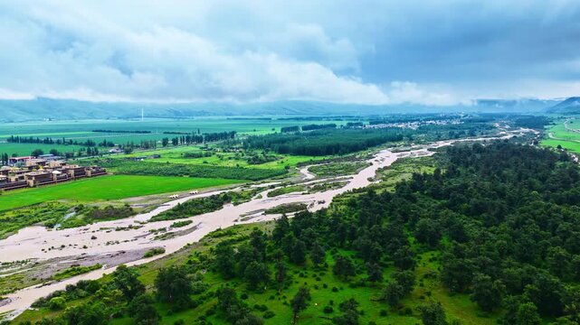 Elevated view of a braided river winding through lush farmland and woodland beneath low clouds in the misty Xinjiang mountains.