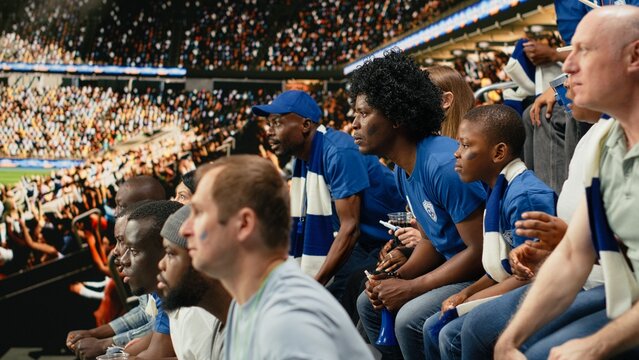 Tense blue team supporters react to a close call during a live soccer match in a crowded stadium while waiting for the final whistle, showing suspense and anticipation across the group.