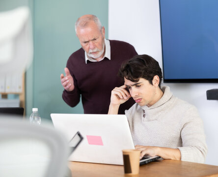 Elderly man sternly scolds young male colleague who working on laptop in office