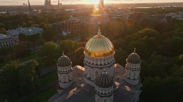 Golden cathedral cupolas at sunset from orbiting drone, Riga, Latvia. Aerial shot circling the cupolas of the orthodox Nativity of Christ Cathedral in Riga, Latvia, as low sun glows over park trees.