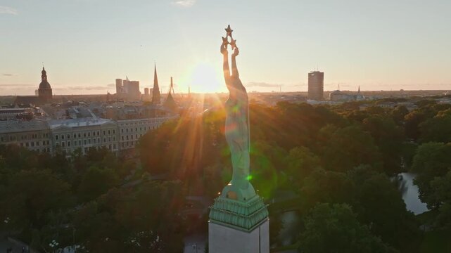 Backlit Freedom Monument Milda from orbiting drone at sunset, Riga. Aerial shot circling the Liberty statue in Riga, Latvia, as sun flares behind the copper figure above city canal.