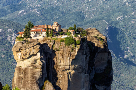 Holy Trinity Monastery perched on a rocky outcrop in the Meteora landscape of Greece, surrounded by dramatic rock formations and mountains.