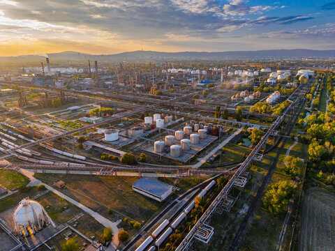 Oil refinery tank farm above parallel rail tracks and pipelines. White cylinders occupy the center, while a spherical vessel enters the lower left beside tank wagons.