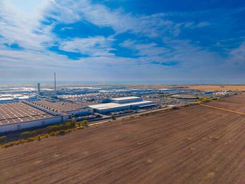 Cultivated field in front of a manufacturing complex. Parallel soil lines fill the foreground beside a road, with factory roofs and parked vehicles beyond.