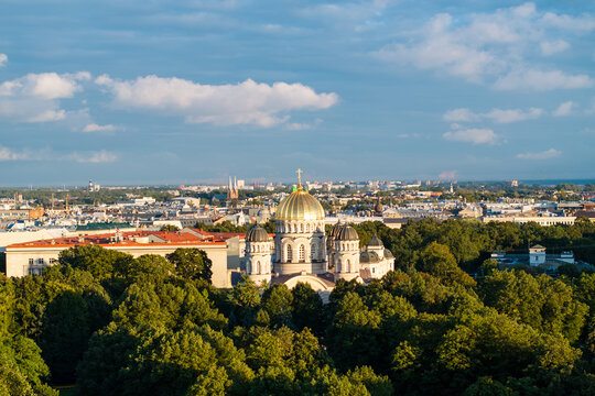 Nativity of Christ Cathedral above park trees under clouds from drone, Riga, Latvia. Aerial view of the gilded domes of the orthodox church rising above green park trees with red roofed building.