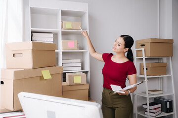 Inventory Management: A woman in a red shirt diligently organizes and inspects boxes stacked on shelves, holding a clipboard for inventory tracking. © Kritdanai