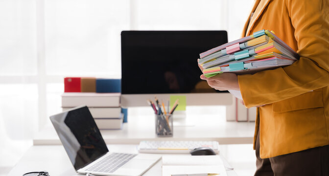 Office Organization: A woman's hands hold a stack of colorful binders and folders on a desk, neatly organized with a laptop and computer, ready for efficient work.