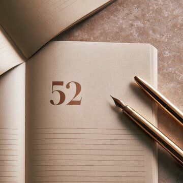 An open journal displays the number 52 on a weekly planner page with a gold pen placed nearby. This setup is meant for blogger annual planning.