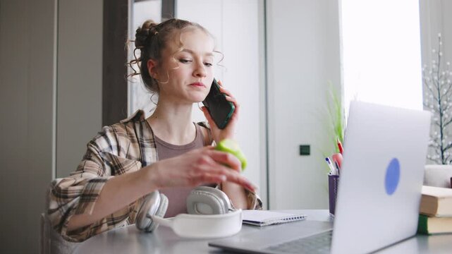 Young woman talks on phone while working on laptop and eating apple in home office setting during day