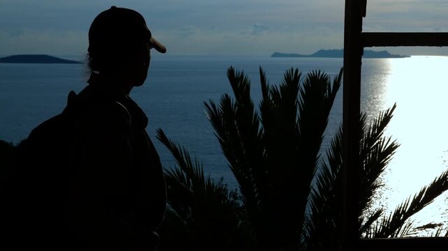 Silhouette of a female hiker admiring the ocean sunset view. Female hiker's silhouette standing on a viewpoint, watching the sun setting over the calm ocean with islands in the distance