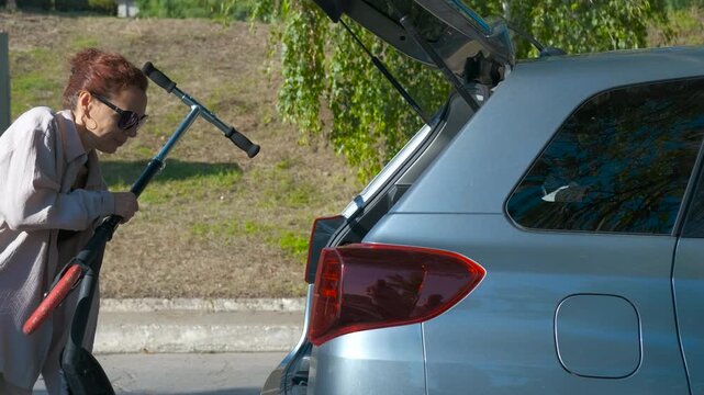 Woman loading folding scooter into car trunk. Red-haired woman loading folding scooter into silver car trunk near urban park, highlighting personal mobility and convenient transportation