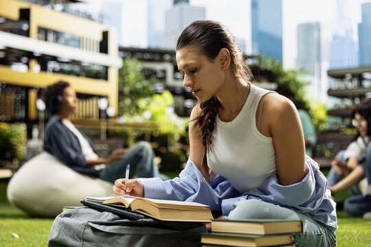 Focused student writes notes beside an open book on a campus lawn, studying alone with stacked books and a backpack during an outdoor study session.