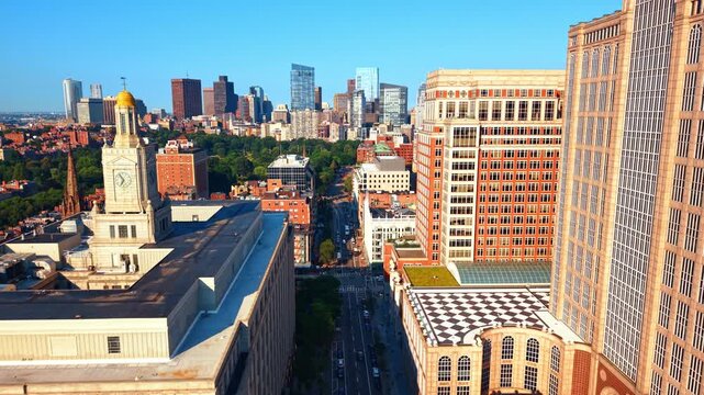 Boston skyline beyond a clock tower and Berkeley Street. An elevated view shows a gold dome, a shaded road and a checkerboard terrace beside brick facades.