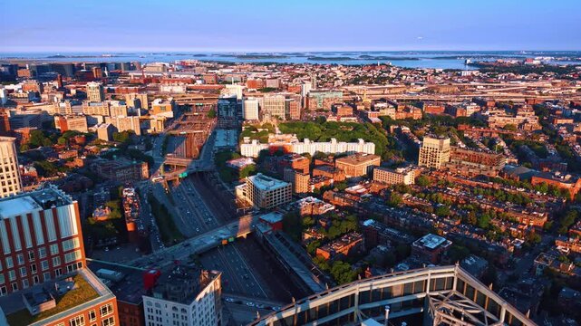 Highway and railway through Boston neighborhoods toward the harbor. Sunlit brick blocks flank parallel transport routes, with bridges, trees and islands beyond.