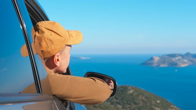 Mature man in car window enjoying sea view during a summer road trip. Middle-aged man with a cap looking out of a car window at a beautiful ocean landscape with islands, enjoying the scenic view