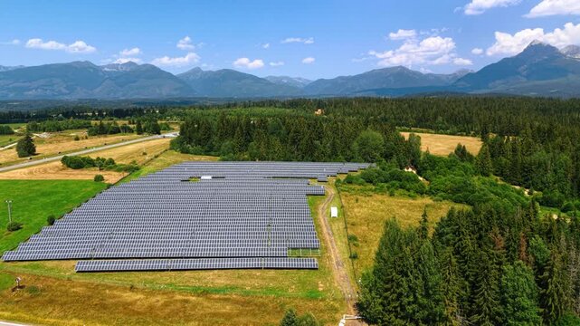 Solar farm bordered by forest beneath the Tatra Mountains. Photovoltaic rows fill a clearing, with conifers on the right and distant peaks beneath clouds.