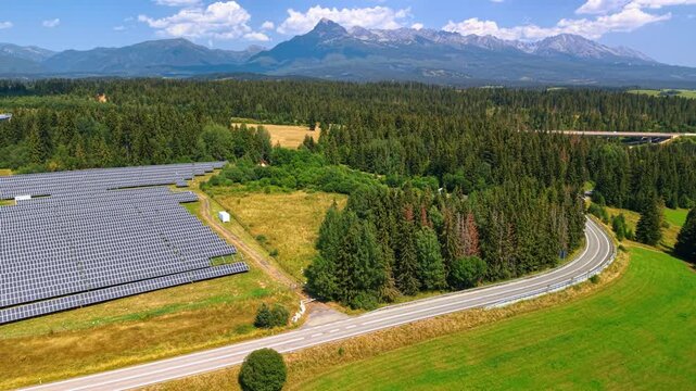 Solar farm beside a road bend beneath the High Tatras. An asphalt curve separates grass from woodland, with panel rows on the left and peaks beyond.