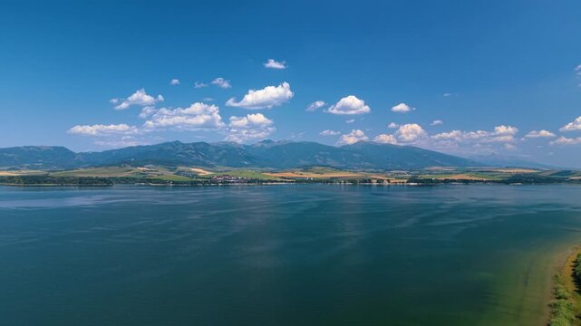 Liptovska Mara reservoir beneath mountain ridges and scattered clouds. Blue water fills the foreground below a distant strip of fields and settlements along the shore.