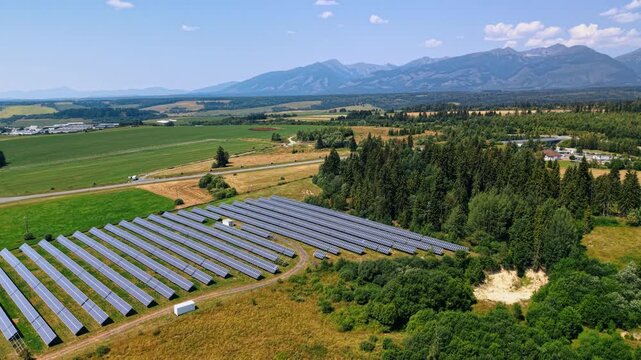 Solar panel rows beside farmland beneath the Tatra Mountains. A photovoltaic array spreads across the lower left, with green fields and mountain slopes behind it.
