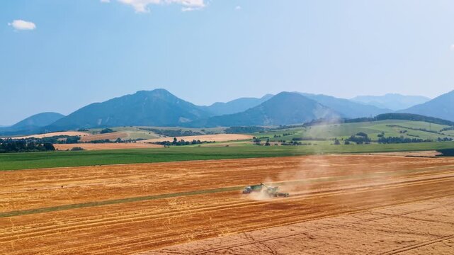 Combine harvester unloading grain beside a tractor in a field. A combine transfers grain into a trailer amid dust, with cut rows and mountain slopes behind it.