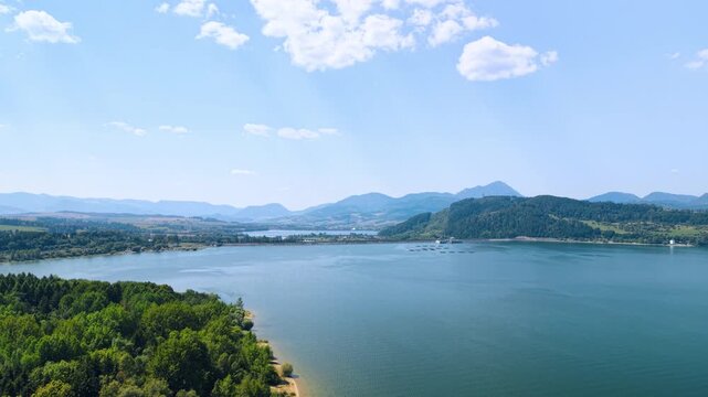 Liptovska Mara reservoir bay between a peninsula and mountain slopes. Green treetops frame the lower left of rippled water, with a wooded hill and distant ridges beyond.