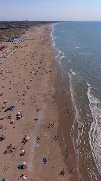 Vertical aerial drone video of Katwijk aan Zee sandy beach, North Sea coast, gentle waves, and distant city skyline in South Holland, Netherlands on sunny summer day