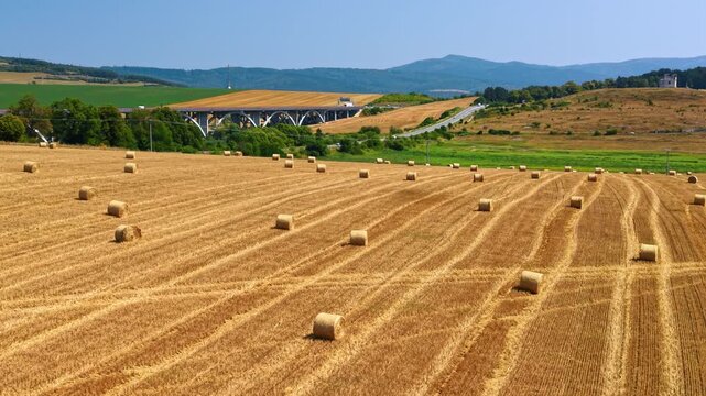 Round straw bales in a harvested field near a viaduct. Rows of golden stubble lead toward concrete bridge arches and a chapel on the hillside.