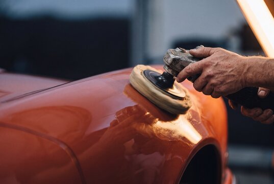 Professional auto detailer polishing orange car body using electric rotary buffing machine in workshop