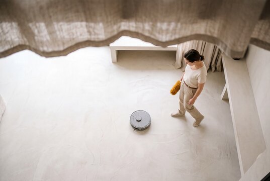 A young woman watches an autonomous robotic vacuum cleaner sweeping the floor in a modern minimalist living room