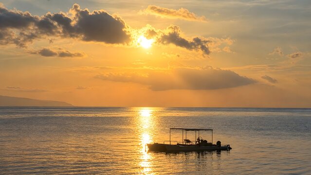 An evocative and dramatic seascape showing a bright golden sun dipping into calm coastal waters, casting a gleaming light path toward a lone fishing boat