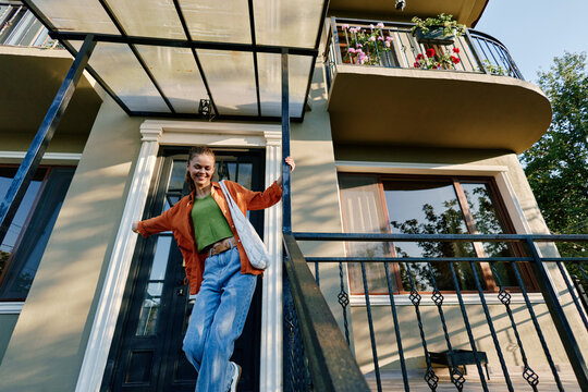 A cheerful young woman in an orange jacket and blue jeans walks down the stairs of a house entrance, smiling with a shoulder bag. Concept of happy lifestyle and new home.