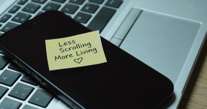 Person places a &ldquo;Less Scrolling More Living&rdquo; sticky note on smartphone, concept of digital detox, reducing screen time and healthier technology habits.