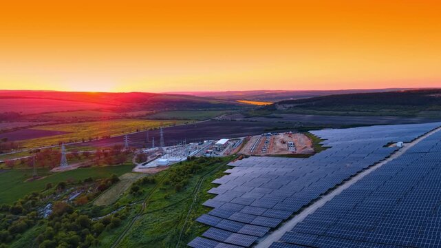Solar farm and electrical substation at sunset. Blue panel rows flank utility equipment and pylons beneath an orange sky and a low sun.