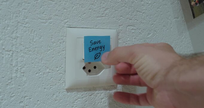 Person places a &ldquo;Save Energy&rdquo; sticky note on a power outlet and switch, concept of energy conservation, reducing electricity consumption and sustainable living.