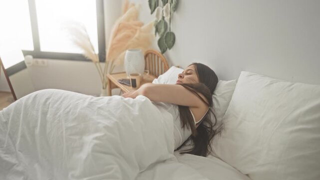 Woman sits up on bed showing bare back and long hair, turning and reaching toward a bright window in a minimalist building interior; serenity.