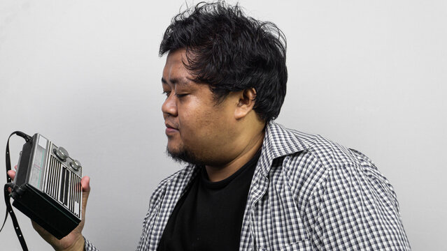 An adult Asian male wearing a checkered shirt over a black shirt holds a portable radio against a plain light gray background. The individual examines the retro broadcasting device