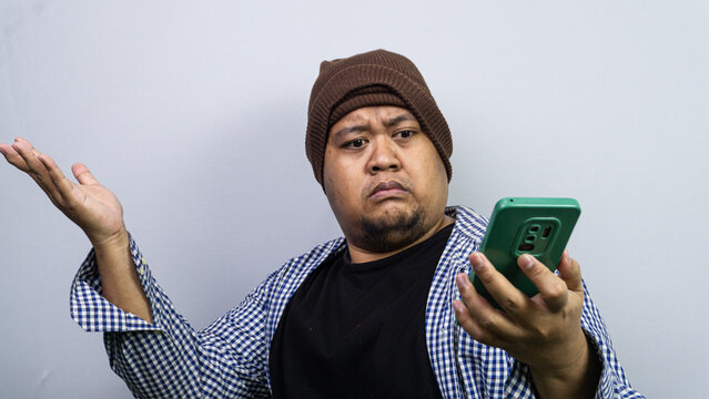A confused Asian male dressed in a checkered shirt and a brown winter beanie gestures questioningly while looking at a green smartphone. The subject raises one hand in disbelief against