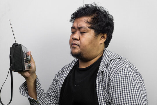 A young Asian male casually dressed in a plaid overshirt inspects a classic analog radio with an extended antenna. The individual stands before a simple white backdrop, projecting