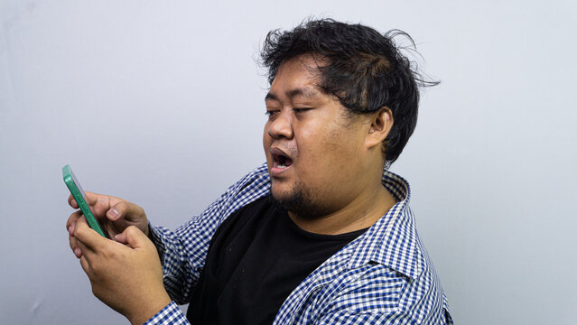A surprised adult Asian male stares at a green smartphone with an open mouth and wide eyes. The amazed individual stands in profile against a plain gray background, reacting to unexpected