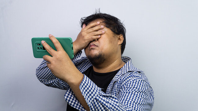 A disappointed adult Asian male covers a hidden face in shame while holding a green smartphone horizontally. The embarrassed individual executes a dramatic facepalm gesture against