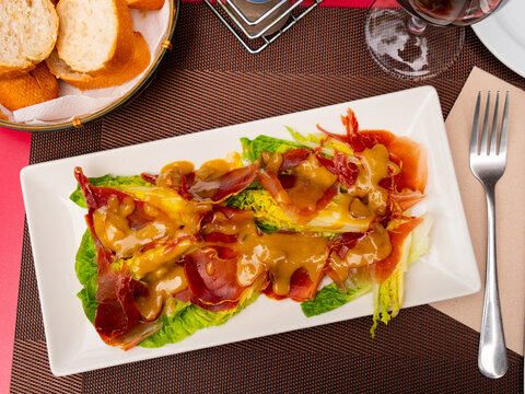 Pork bacon ham slices are baked with young, crisp cogollos lechuga hearts and seasoned with mustard sauce. Meat and vegetable appetizer, set against brown background