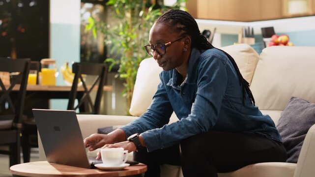 Black woman checks the hour on her wristwatch for time management, anxious about networking tasks and schedule. Young adult digital nomad enjoys domestic life and punctuality at home.
