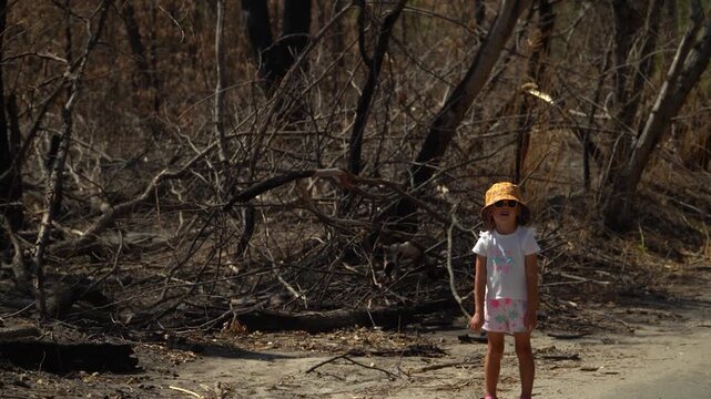 spring day, filming forest, remains burnt bushes trees, dried-up river background, natural disaster, absence animals birds, girl yellow Panama hat stands against backdrop burnt forest bored look