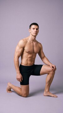 Fit muscular man demonstrating powerful pose kneeling on one knee against a plain purple-gray background