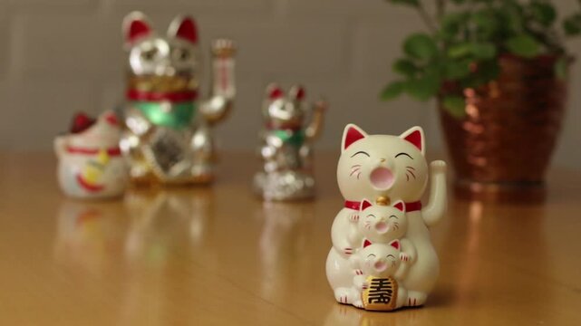 Video de gato de la suerte Maneki Neko moviendo la pata, al fondo 3 gatos de la suerte y una planta verde fuera de foco